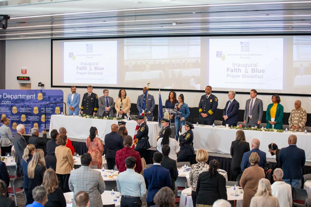 Attendees and head table guests stand as the APD honor guard presents the colors and a young singer holds a microphone at the lectern during the inaugural Faith & Blue Prayer Breakfast.