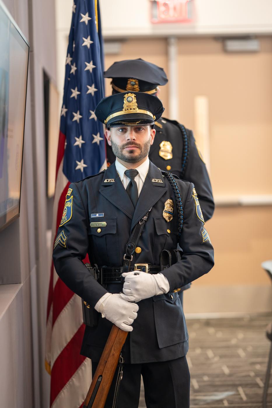 An APD honor guard member in dress uniform and white gloves holds a ceremonial rifle, with another member and an American flag behind him.
