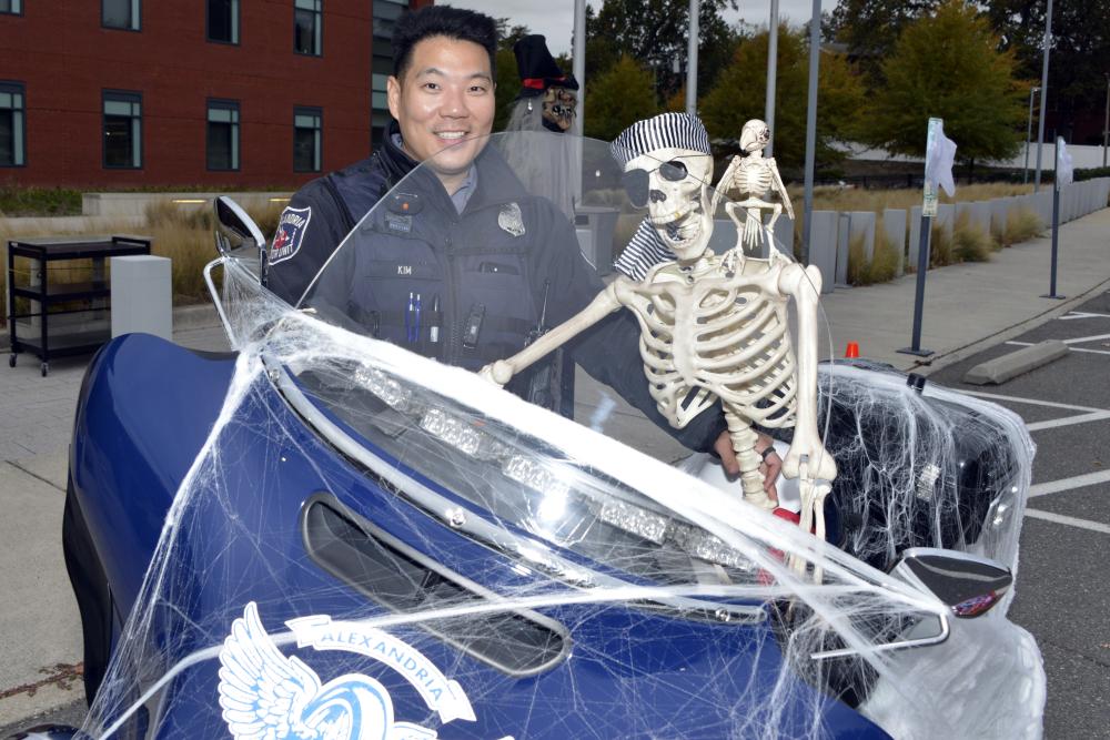 A smiling police officer stands behind a police motorcycle draped in artificial cobwebs, with a skeleton seated beside him.