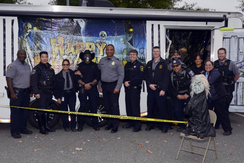 A group of police personnel poses in front of a Halloween backdrop with skeleton decorations and yellow caution tape.