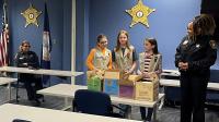 Three Girl Scout Cadettes delivering four cartons of cookies to two deputies in a roll call room