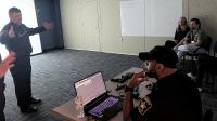 Uniformed Sheriff's Office commander speaking to three civilians in a dark room with a white screen. Another deputy operates at laptop.
