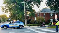 Blue and gold sheriff's cruiser with blue and red lights flashing escorting several children riding bicycles down a street, with a crossing guard visible on the right at a crosswalk