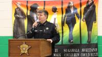 Sheriff in blue uniform standing at podium and speaking into a microphone with a banner of Harriet Tubman, Rosa Parks, Ruby Bridges, Kamala Harris and Ketanji Brown Jackson in the background