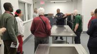 Deputy in blue uniform holding meal service tray of food inside institutional kitchen with several community members present