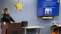 Sergeant in a blue uniform standing a podium and looking toward a screen for a presentation on the booking process