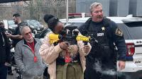 Civilian firing a pepperball training launcher under the supervision of a Sheriff's sergeant in a blue uniform.