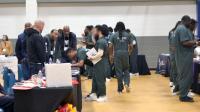 Several inmates in green jumpsuits speaking with exhibitors at tables in a gym
