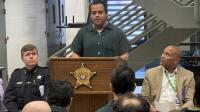 Inmate in a green jumpsuit speaking at a podium with a Sheriff's star on the front