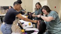 Hair braiding instructor providing a student-inmate with guidance on braiding hair on a mannequin head with four other student-inmates looking and working on braiding their mannequin heads
