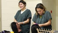 Two inmates in green uniforms braiding the hair on two mannequin heads