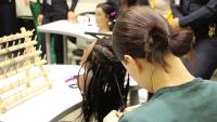 View from behind of an student wearing a green prisoner shirt and braiding hair on a mannequin head