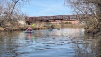 Three kayakers one after the other on the river with paddles, just went under a bridge at Four Mile Run Park