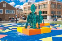 Site See Installation with human figures from torso up in green metal with holes and an orange and yellow square foundation. Cement is also painted blue and geometrical shapes of yellow and pink
