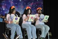 Three tween girls wearing white shirts and jeans sitting on chairs holding pieces of paper with flowers on them and microphones