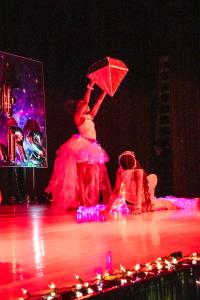 Performer in a dress standing while holding a giant diamond with another performer posing on the floor. Red light shining on both of them