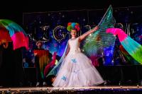 Performer in the middle in a white dress with one arm up holding a translucent wing and wearing a colorful headwrap