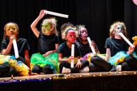 Child performers sitting on the stage dressed in black with colorful painted faces while holding white tubes