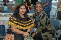 woman with black and yellow dress and woman with green and black sweater sit next to each other and smile at camera
