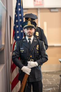 An APD honor guard member in dress uniform and white gloves holds a ceremonial rifle, with another member and an American flag behind him.