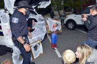 An officer wearing a black hat and skull face covering stands beside a cobweb-covered vehicle as an adult holds up a phone to take a photo.
