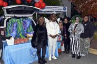 Six adults, several wearing Halloween costumes, pose beside an open hatch decorated with a colorful backdrop and red and black balloons.