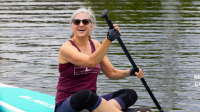 Woman in a maroon tank top, wearing black sunglasses, sitting on a blue paddleboard while holding a paddle and smiling wide towards a person who's blurred on the left side of the image