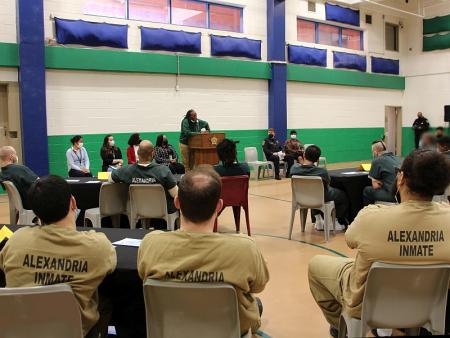 speaker at podium with Sheriff and civilians sitting on both sides facing an audiences of inmates in tan and green jumpsuits sitting at tables