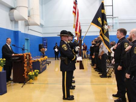 deputies in uniform looking at flag, honor guard holding flags and civilian singing at podium