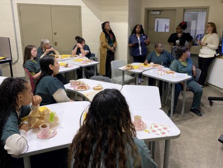Eight women in green jumpsuits sitting at tables with tea and breakfast foods listening to a women in a blue jacket speak. Three other women are also standing and listening to the speaker.