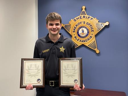 Deputy in blue uniform holding two award plaques and standing in front of a large gold sheriff's star on a blue wall