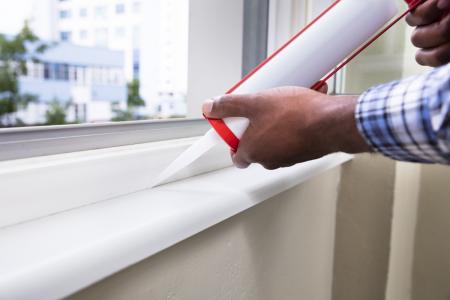 A pair of hands hold a caulk gun to apply caulk along a window frame.