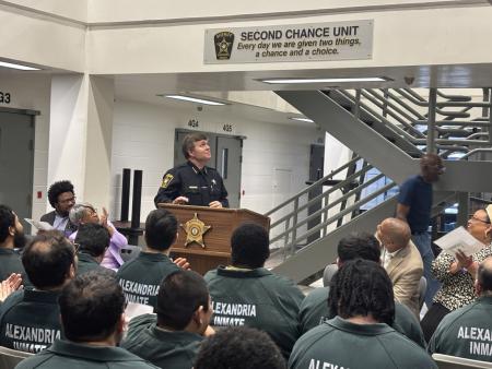 Sheriff standing at podium, looking up and smiling at sign that says Second Chance Unit. Several seated inmates wearing green jumpsuits and visible from the back.