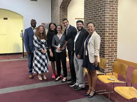 Members of the Alexandria City Council surround Tamara Jovovic, who is holding the plaque she was awarded. Ms. Jovovic is a white woman with brown hair and glasses.