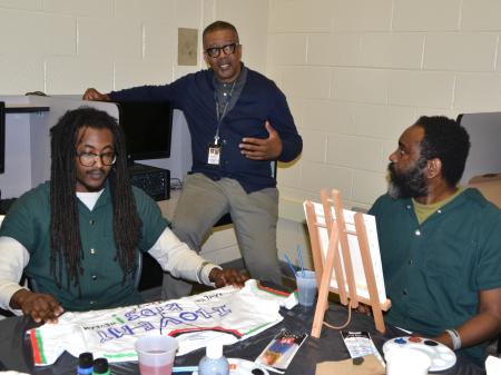 Reentry counselor speaking with two men in green jumpsuits who are painting, one painting a t-shirt and the other a small canvas on an easel