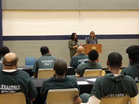 Several inmates in green jumpsuits seated in a gymnasium visible from behind with two guest speakers at a podium in the background