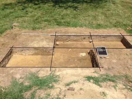 Four square excavation units are shown in a field. Each unit has straight vertical sides and a flat soil floor. Lines are projected across the units digitally to show the excavation grid.
