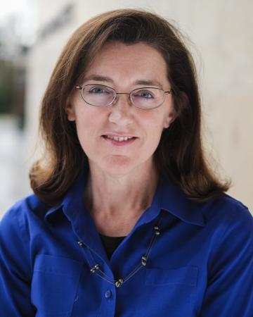 A woman wearing glasses and a blue shirt smiles at the camera in a professional headshot. 