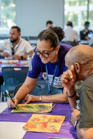 This image shows two people engaged in an art activity at a table covered with a purple cloth.