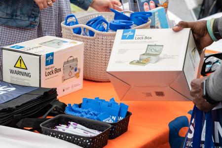 A close-up shot shows a person's hands holding a white box for a Medline Medication Lock Box above an orange-covered table. The table is filled with community resources, including more lock boxes, a basket of blue water bottles, pens, and tote bags labeled for suicide prevention. In the background, another individual is seen browsing the items on display.