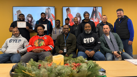 Fatherhood Program Group Photo. A group of approximately 13 men poses for a photo in a room with yellow walls. Several men are wearing sweatshirts with the "CYEP" logo, and two men in the back row hold up certificates. In the foreground, a festive evergreen centerpiece with a candle sits on a wooden table.