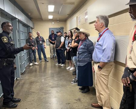 Deputy in a blue uniform giving a tour to several civilians with prisoner cells visible on the left side