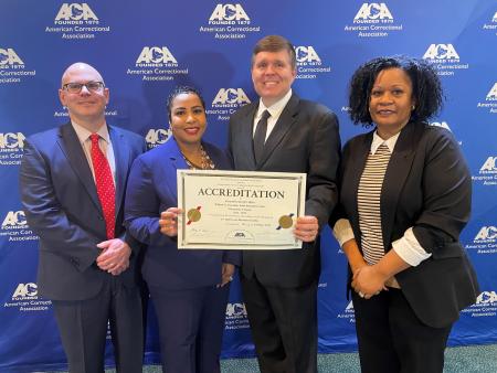 Four corrections executives in business attire standing next to each other with two in the center holding a certificate