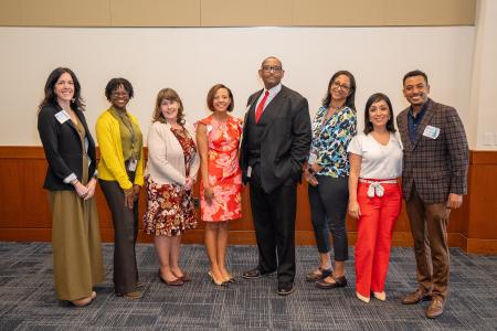 The image shows a group of eight professionally dressed individuals standing in a line, posing for a photo at an indoor event. They are standing against a neutral-toned wall with wood paneling on the lower half and a dark blue patterned carpet.