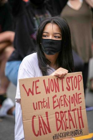 A woman in a cloth pandemic mask holds a brown cardboard side reading, “We won’t stop until everyone can breath (sic). #Blacklivesmatter.”