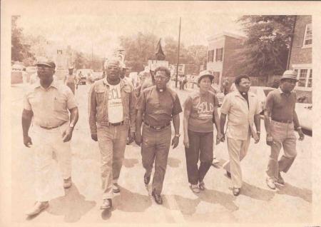 Faith leaders march in the SCLC protest, including men in SCLC t-shirts and a priest in a collar.