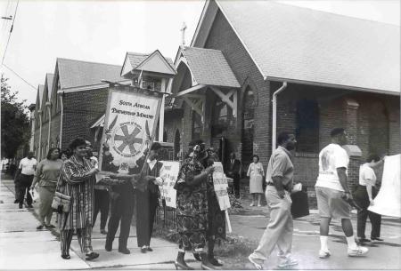 A procession marking the end of apartheid passes a brick church in this black-and-white photograph. A large sign reads “South African Partnership Ministry.”