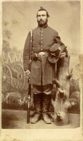 Private Francis E. Brainerd holds his rifle and his hat, leaning on a tree stump with a painted scene of a Civil War encampment behind him in this studio photograph.