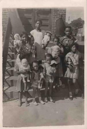 A group of Black children pose with their dolls on the steps of a brick building.
