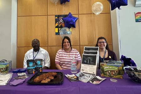 Three staff members sit behind a purple information table decorated with balloons during a Purple Up Day event. The table displays brochures, military family resources, giveaways, cookies, and support materials for Service Members, Veterans, and their Families (SMVF).